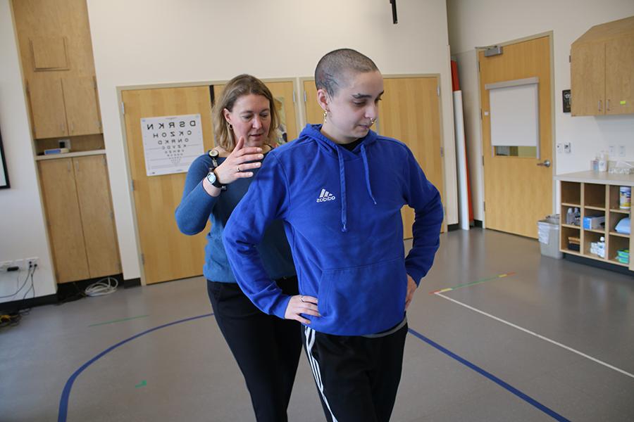 A therapist stands behind a patient as she practices balancing.